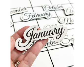 Puede incluir: Azulejos de calendario de madera blanca con letras cursivas negras para cada mes del año. El azulejo de enero se sostiene en una mano.