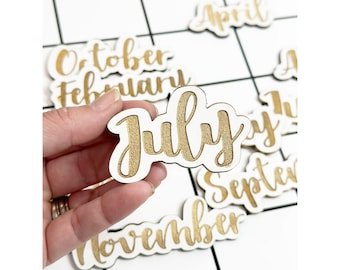 Puede incluir: Azulejos de calendario de madera blanca con letras doradas que deletrean los meses del año. El azulejo de julio se sostiene en una mano.
