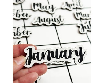 May include: White wooden calendar tiles with black lettering spelling out the months of the year. The tile for January is being held in a hand.