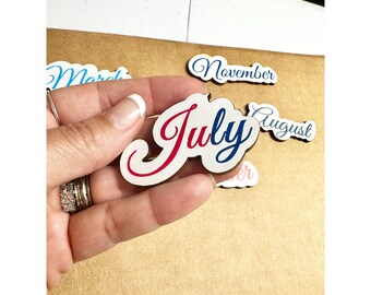 May include: A white wooden cutout of the word "July" in red and blue cursive lettering. The cutout is being held in a hand.