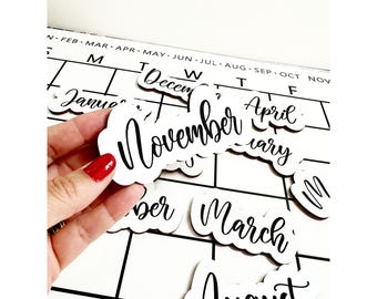 May include: A hand holding a white wooden cutout of the month "November" with black cursive lettering. Other month cutouts are scattered on a calendar grid, including "January", "March", and "August".