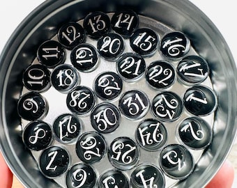 May include: A set of 31 black and white glass number tiles in a silver tin. The numbers are in a cursive font and range from 1 to 31.