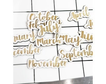 May include: Gold-colored month stickers on a white background. The months are January, February, March, April, May, June, July, August, September, October, November, and December.