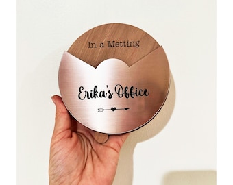 Könnte beinhalten: Ein rundes Holzschild mit einer metallischen roségoldenen Auflage. Das Holz ist mit dem Text In a Metting graviert. Der roségoldene Bereich zeigt den schwarzen Text Erika's Office mit einem kleinen Pfeil und Herzsymbol unter dem Namen.