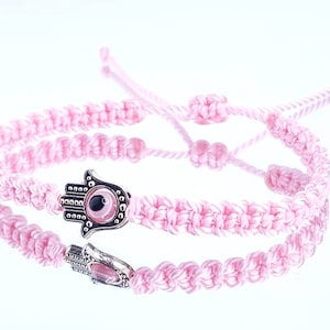 Puede incluir: Pulsera trenzada rosa con un dije de plata que representa una mano de Hamsa con un ojo turco en el centro.