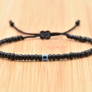 May include: Black beaded bracelet with an adjustable cord closure. The bracelet features a single blue and white evil eye bead in the center. The beads are small and round, and the cord is knotted to adjust the size.
