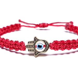 May include: Red braided bracelet featuring a Hamsa hand charm with an evil eye design. The bracelet is adjustable and made of red cord. The Hamsa charm is a bronze color with a blue and white eye in the center.