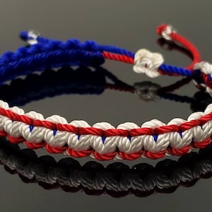 May include: A red, white, and blue braided friendship bracelet. The bracelet is made with three strands of cord, and the colors are arranged in a pattern that resembles the American flag.