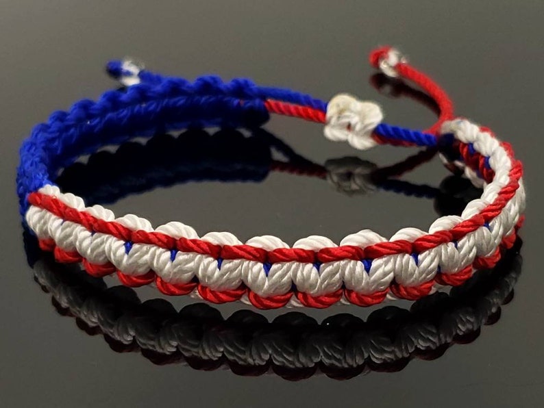 May include: A red, white, and blue braided friendship bracelet. The bracelet is made with three strands of cord, and the colors are arranged in a pattern that resembles the American flag.