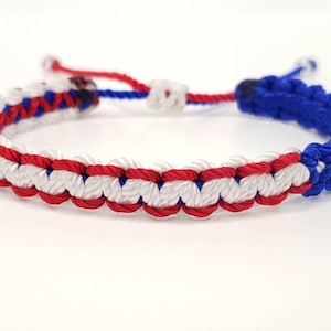 May include: A red, white, and blue braided friendship bracelet. The bracelet is made with a knotting technique and has a simple design.