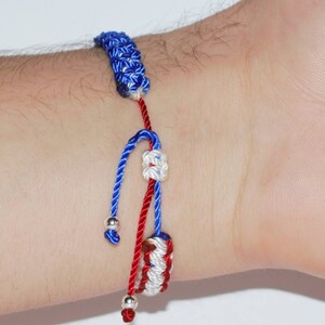 May include: A red, white, and blue braided bracelet with silver beads. The bracelet is tied in a knot and has a decorative knot at the end.