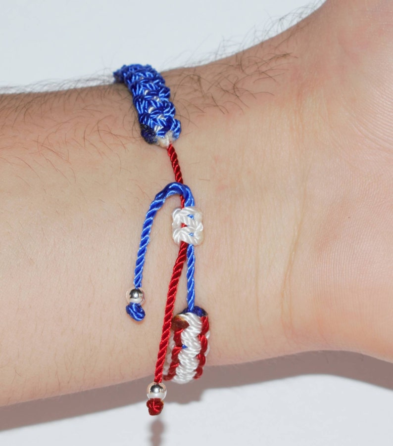 May include: A red, white, and blue braided bracelet with silver beads. The bracelet is tied in a knot and has a decorative knot at the end.