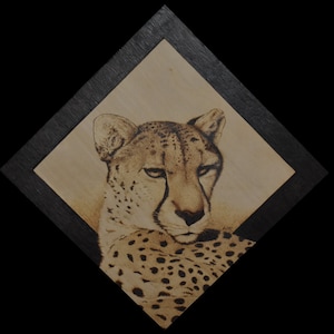 K&ouml;nnte beinhalten: Eine Nahaufnahme eines Holzbrandbildes eines Gepardenkopfes. Der Gepard schaut nach rechts im Bild, mit leicht geneigtem Kopf. Das Fell des Geparden ist hellbraun mit schwarzen Flecken.