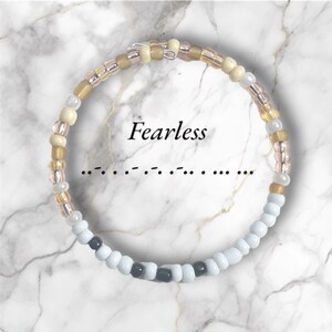 May include: A beaded bracelet with white, black, gold, and pink beads. The bracelet has the word "Fearless" written on it.