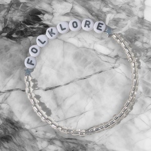 May include: White beaded bracelet with the word "FOLKLORE" spelled out in black letters. The bracelet is on a marble background.