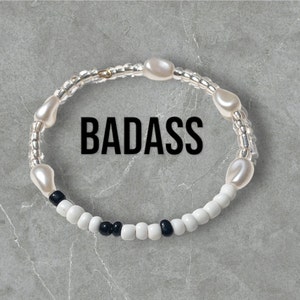 May include: A white beaded bracelet with black beads and white pearl-like beads. The bracelet has the word "BADASS" written on it.