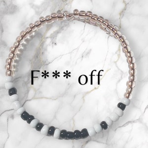 May include: A black and white beaded bracelet with a gold-toned metal clasp. The bracelet has the text "F*** off" written on it.