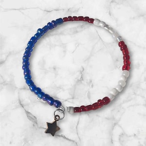 May include: A beaded bracelet with red, white, and blue beads, with a gold star charm. The bracelet is on a white marble surface.