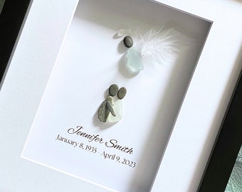 May include: A framed piece of art featuring a white feather, a blue glass stone, and two gray stones arranged to look like a couple. The text "Jennifer Smith January 8, 1935 - April 9, 2023" is written below the stones.