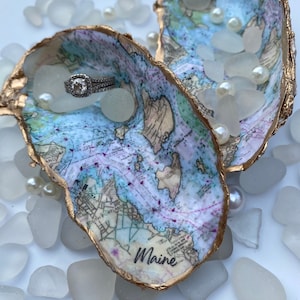 May include: Two gold-rimmed oyster shells with a nautical map design. The shells are filled with white sea glass and pearls. One shell has a diamond engagement ring inside. The shell on the right has the word "Maine" written on it.