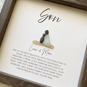 May include: A framed artwork with a dark gray wooden frame. The artwork features the word "Son" in cursive, and a pebble art depiction of a parent and child. The text reads "Liam & Mom" and a heartfelt message.