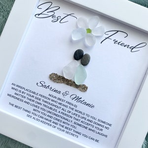 May include: White frame with a white background and black text. The text reads "Best Friend" and "Sabrina & Melanie". The text continues with a definition of a best friend. The frame contains a flower made of white sea glass and gray stones.