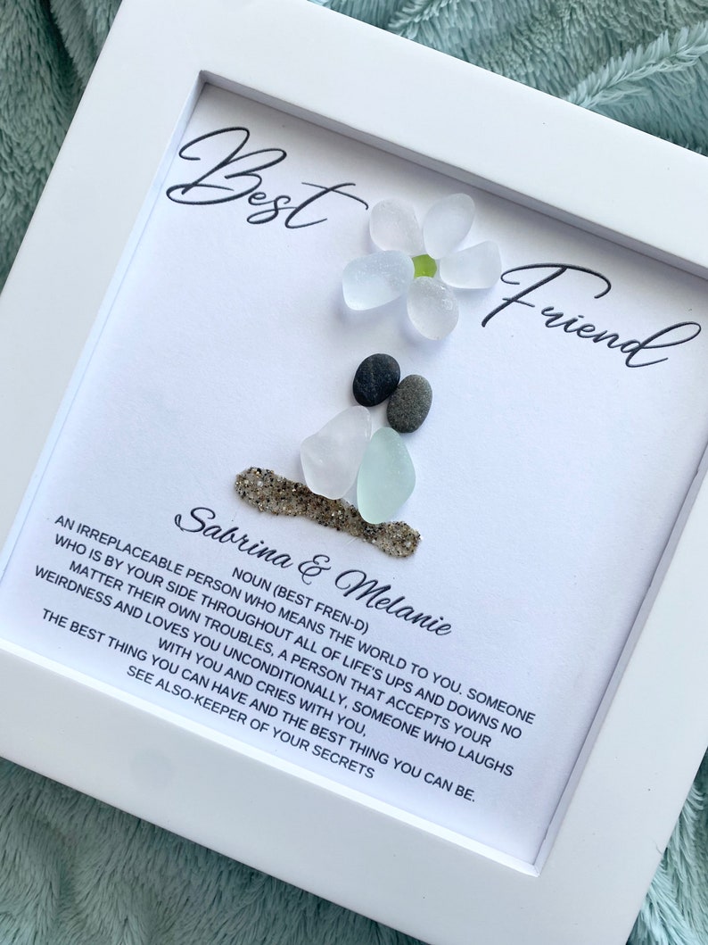May include: White frame with a white background and black text. The text reads "Best Friend" and "Sabrina & Melanie". The text continues with a definition of a best friend. The frame contains a flower made of white sea glass and gray stones.