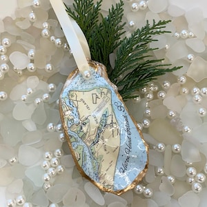 May include: Oyster shell Christmas ornament featuring a map of Hilton Head Island, SC. The shell has a gold-painted edge and a light blue interior. A white ribbon and sprig of greenery are attached. The ornament is surrounded by pearls and sea glass.