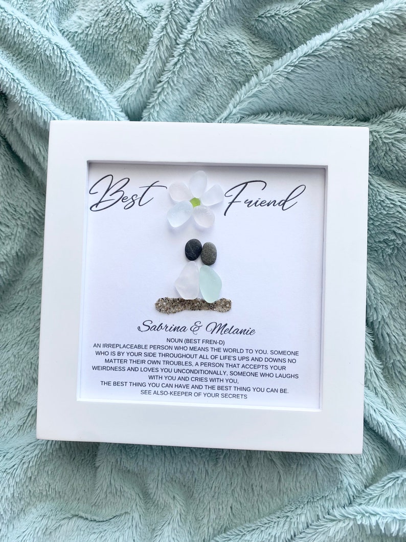May include: White framed artwork with a sea glass flower and two pebbles. The text reads "Best Friend" and "Sabrina & Melanie" with a definition of the word "friend".