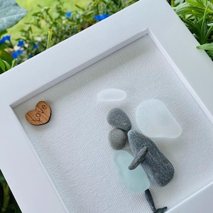 May include: A framed artwork featuring a pebble art angel with a blue and white glass wing. The angel is holding a heart with the word "Love" inscribed on it.