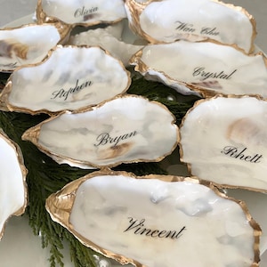 May include: A collection of oyster shells with gold-painted edges, each inscribed with a name in elegant black script. The shells are arranged on a white plate with greenery, creating a sophisticated table setting.