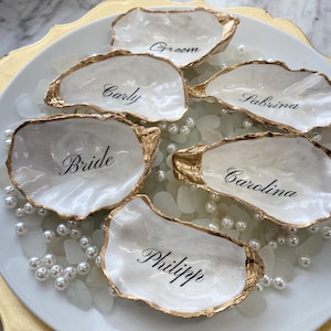 May include: Six white oyster shells with gold-painted edges, arranged on a white plate with pearls. Each shell has a name written in black script: Bride, Groom, Carly, Sabrina, Carolina, and Philipp. A decorative wedding or event theme.