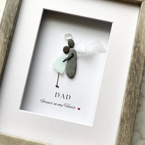 May include: Framed artwork with pebble art of two figures embracing, with a white feather wing. The text reads "DAD Forever in my Heart" in black script, with a small red heart. The frame is a light grey colour.