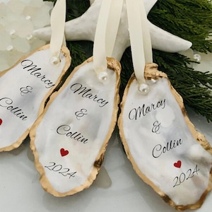 May include: Three white oyster shell ornaments with gold trim. Each ornament has the names "Marcy & Collin" and the year "2024" written on them. The ornaments are tied with white ribbon.