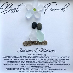 May include: A white card with the words "Best Friend" written in black cursive. The card features a flower made of white sea glass and two figures made of sea glass and stones. The text below the image reads "Sabrina & Melanie" and includes a definition of the word "friend".