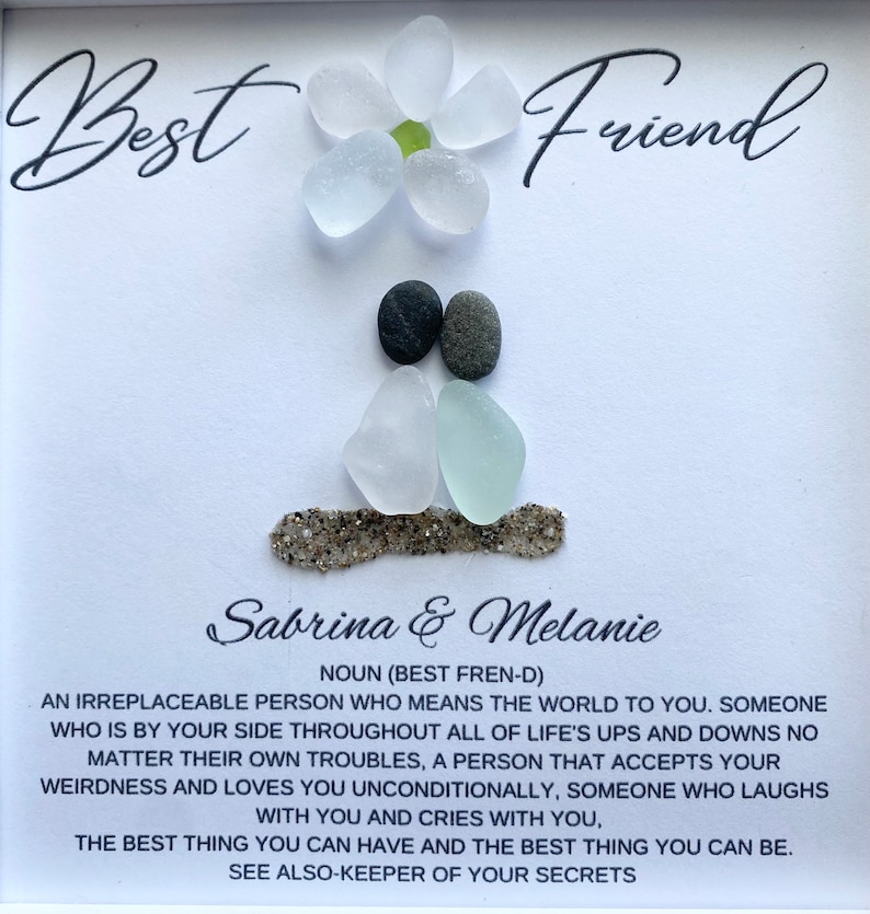 May include: A white card with the words "Best Friend" written in black cursive. The card features a flower made of white sea glass and two figures made of sea glass and stones. The text below the image reads "Sabrina & Melanie" and includes a definition of the word "friend".
