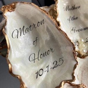 May include: Two white oyster shells with gold trim, each with a handwritten inscription. The left shell reads "Matron of Honor 10-1-25". The right shell reads "Mother of the Groom 10-1-25".