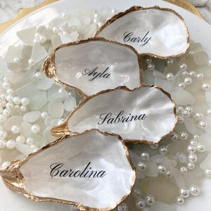 May include: Four white oyster shells with gold-painted edges, each inscribed with a name in elegant black script: Carly, Ayla, Sabrina, and Carolina. The shells rest on a white plate with pearls and sea glass accents, creating a coastal theme.