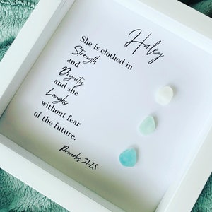 K&ouml;nnte beinhalten: Ein wei&szlig;er gerahmter Druck mit dem Text "She is clothed in Strength and Dignity and she Laughs without fear of the future. Proverbs 31:25" und dem Namen "Haley" in Kursivschrift. Auf dem Druck befinden sich drei kleine blaue und wei&szlig;e Glassteine.