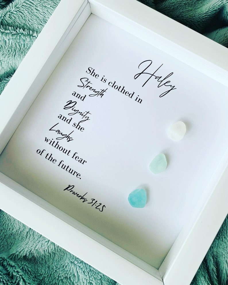 K&ouml;nnte beinhalten: Ein wei&szlig;er gerahmter Druck mit dem Text "She is clothed in Strength and Dignity and she Laughs without fear of the future. Proverbs 31:25" und dem Namen "Haley" in Kursivschrift. Auf dem Druck befinden sich drei kleine blaue und wei&szlig;e Glassteine.