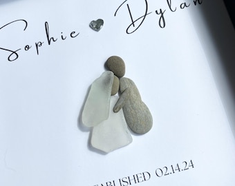 May include: A framed artwork featuring a couple depicted with stones and sea glass. The names "Sophie" and "Dylan" are written in cursive with a small silver heart between them. The text "ESTABLISHED 02.14.24" is written below the couple.