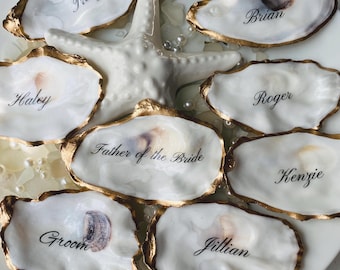 May include: A collection of white oyster shells with gold-painted edges, each inscribed with a name in elegant black script. A white starfish and scattered pearls accent the shells, arranged on a white plate, ideal for wedding place settings.
