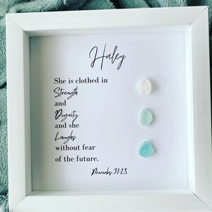 K&ouml;nnte beinhalten: Ein wei&szlig;er gerahmter Druck mit dem Namen "Haley" oben und einem Zitat &uuml;ber St&auml;rke, W&uuml;rde und Lachen. Das Zitat stammt aus Sprichw&ouml;rter 31:25. Drei kleine St&uuml;cke Meerglas sind vertikal auf der rechten Seite des Drucks angeordnet.
