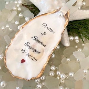 May include: An oyster shell ornament with gold trim, inscribed with "Willow & Stephen Engaged 10/24/25" and a red heart. The ornament is adorned with a pearl and ribbon, resting on a bed of pearls and sea glass, with a white starfish in the background.