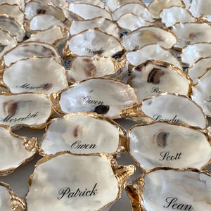 May include: A collection of oyster shells with gold-painted edges, each inscribed with a name in elegant black script. The shells are arranged closely together, creating a textured surface. The shells are white with brown and gold accents.