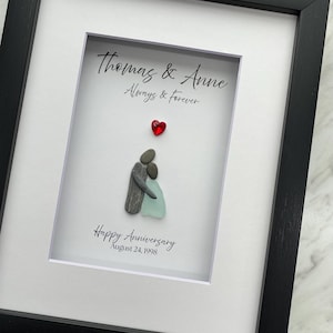 May include: Framed artwork with a black frame and white matting. The artwork features a pebble art depiction of a couple embracing, with a red heart above them. Text reads "Thomas & Anne Always & Forever" and "Happy Anniversary August 24, 1998."