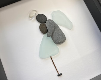 Könnte beinhalten: Ein gerahmtes Kunstwerk mit einem Engel aus Meeresglas. Der Engel besteht aus verschiedenen Formen von Meeresglas und Kieselsteinen, mit einem hellblauen Kleid, grauen Flügeln und einem dunkelbraunen Stiel. Das Kunstwerk ist vor einem weißen Hintergrund in einem schwarzen Rahmen.
