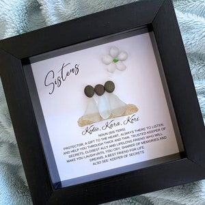 May include: A framed art piece with the word "Sisters" in elegant script. The artwork features three figures made of pebbles and sea glass, with the names Kylie, Kara, and Kori. A definition of sisters is included in the text.