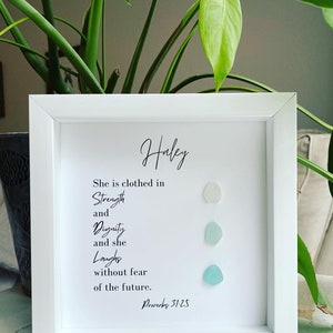 K&ouml;nnte beinhalten: Ein wei&szlig;er gerahmter Druck mit dem Namen "Haley" oben. Der Druck zeigt ein Zitat &uuml;ber St&auml;rke, W&uuml;rde und Lachen mit dem Bibelvers "Spr&uuml;che 31:25". Drei kleine, hellblaue Glasscherben sind vertikal auf der rechten Seite des Drucks angeordnet.