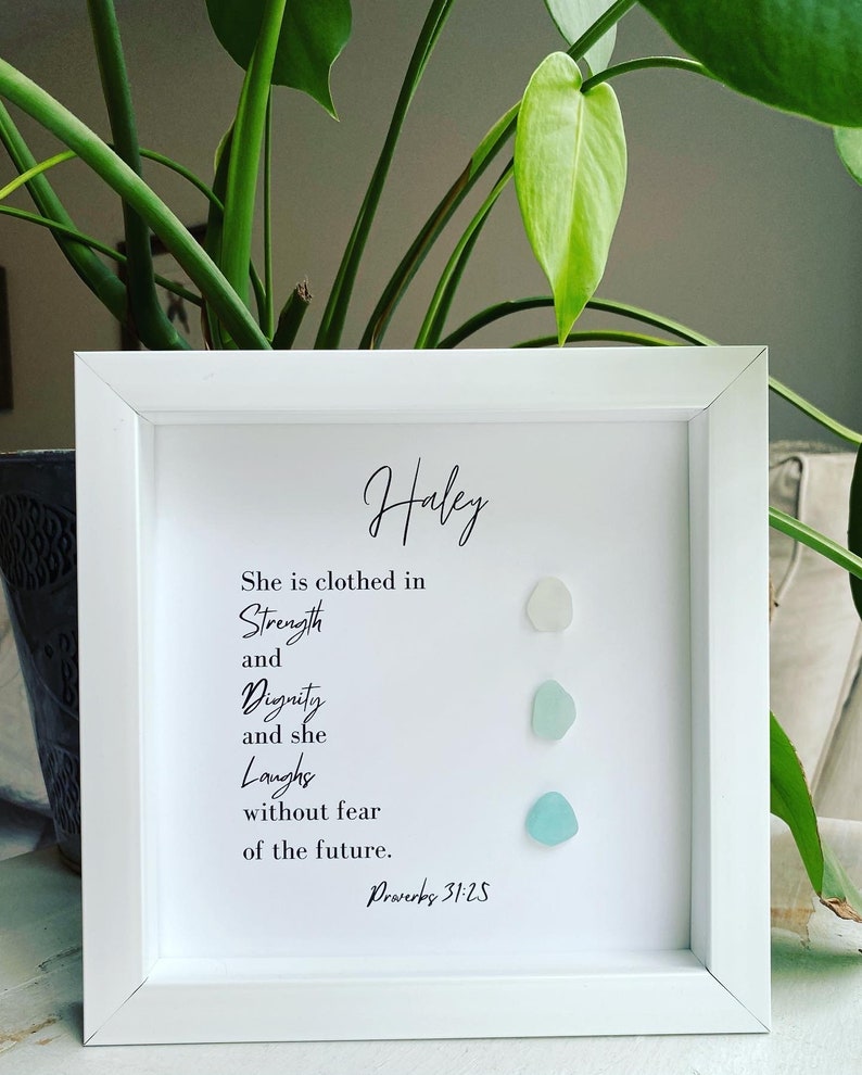 K&ouml;nnte beinhalten: Ein wei&szlig;er gerahmter Druck mit dem Namen "Haley" oben. Der Druck zeigt ein Zitat &uuml;ber St&auml;rke, W&uuml;rde und Lachen mit dem Bibelvers "Spr&uuml;che 31:25". Drei kleine, hellblaue Glasscherben sind vertikal auf der rechten Seite des Drucks angeordnet.
